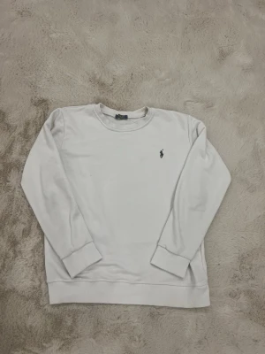 Vit sweatshirt från Polo Ralph Lauren - Klassisk vit sweatshirt från Polo Ralph Lauren med rund halsringning och broderad logga på bröstet. Tröjan har långa ärmar och ribbade muddar vid ärmslut och nederkant. Perfekt för en clean och stilren look.