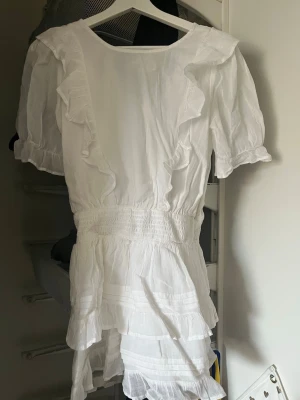 Vit volangklänning med kort ärm - Supersöt vit klänning med volangdetaljer och korta puffärmar. Klänningen har smock i midjan för en snygg passform och dubbla volanger i kjolen. Materialet ser ut att vara lätt och luftigt, perfekt för varma dagar. Baksidan har en v-ringning med knytband.