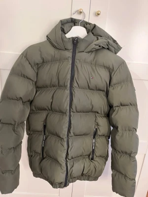 Olivgrön pufferjacka från Tommy Hilfiger - Snygg olivgrön pufferjacka från Tommy Hilfiger med huva och dragkedja framtill. Jackan har två fickor med dragkedja och diskret logga på bröstet. Perfekt för kalla dagar med sin vadderade design och sportiga look.