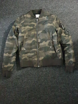 Camo bomberjacka från KappAhl - Snygg bomberjacka i camomönster från KappAhl. Jackan har dragkedja framtill, två fickor med tryckknapp och en ärmficka med dragkedja. Muddar vid ärmslut, hals och nederkant. Perfekt för dig som gillar streetstyle.