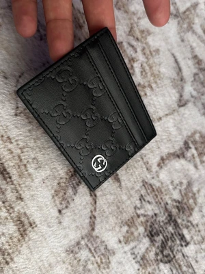 Svart kortfodral från Gucci - Snyggt svart kortfodral från Gucci i läder med präglat GG-mönster och silverfärgad logga på framsidan. Fodralet har flera fack för kort och är stilrent och kompakt, perfekt för att hålla koll på dina viktigaste kort.