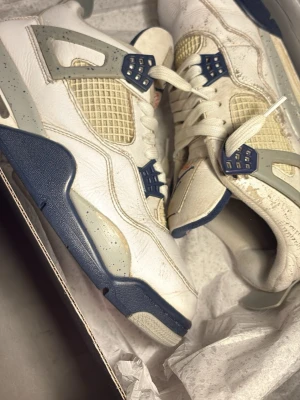 Air Jordan 4 Retro Vit/Blå - Säljer ett par Air Jordan 4 Retro sneakers i vitt med blå och grå detaljer. Skorna har meshpaneler, klassiska plastvingar och tjock sula med speckle-mönster. Ikonisk Jumpman-logga på plösen och häl. Dem är lite slitna på sitan men det är inget man märker så mycket hade jag sakt. Skick utseende mässigt från nya hade jag kanske sakt 6/10 skorna är mellan bra och acceptabelt skick för jag tvättar dem väldigt noga när jag kan.