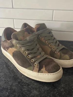 Camo sneakers i mocka - Snygga sneakers med camouflagemönster i olika gröna och bruna toner från Sandays footwear. Ovandelen är i mocka och skorna har gröna skosnören samt guldfärgade öljetter. Vit sula och lågt skaft ger en clean look som passar till det mesta. Perfekt för vår och sommaren. Priset är ej hugget i sten 