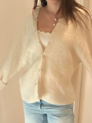 Krämvit oversized kofta med knappar - Mysig krämvit kofta med v-ringning och pärlemorskimrande knappar framtill. Koftan har en oversized passform, långa ärmar och är stickad i ett mjukt material. Perfekt att slänga över ett linne eller t-shirt för en chill look.