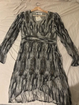 Mönstrad omlottklänning från SOFIE SCHNOOR - Klänning från Sofie Schnoor i storlek XS (Passar S). Mönstrad i grå/svarta toner med lätt skimrande tyg, djup V-ringning och markerad midja. Långärmad modell med fint fall. I mycket fint skick, prislapp kvar, aldrig använd. Originalpris:1499kr