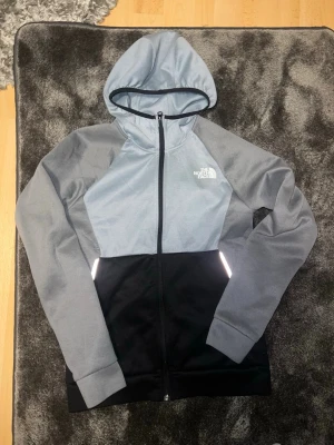 Grå och svart hoodie från The North Face - Snygg hoodie från The North Face i grått och svart med hel dragkedja och huva. Loggan är tryckt i vitt på bröstet. Hoodien har långa ärmar och praktiska fickor framtill. Perfekt för en chill och sportig look.
