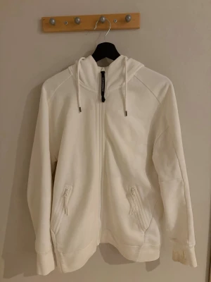 Vit hoodie med glasögon i huvan - Säljer en vit zip hoodie från C.P. Company I jättefint skick. Hoodien har dragkedja framtill, två fickor med dragkedja och glasögon i huvan som man kan knäppa upp och ner.