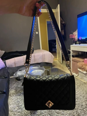 Svart quiltad handväska med kedja - Snygg svart handväska med quiltat mönster och guldfärgad kedjerem. Väskan har en elegant fyrkantig form och ett dekorativt guldfärgat spänne framtill. Perfekt accessoar för att lyfta din outfit.