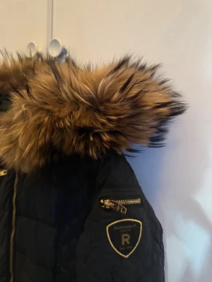 Svart dunjacka med päls från Rockandblue - Snygg svart dunjacka från Rockandblue med quiltad design och guldiga dragkedjor. Jackan har en stor huva med fluffig brun äkta päls och ett emblem på ärmen. Perfekt för kalla vinterdagar och riktigt stilren look.