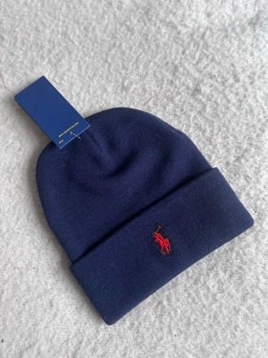 Mörkblå mössa från Polo Ralph Lauren - Snygg mörkblå mössa från Polo Ralph Lauren med klassisk uppvikt kant och röd broderad logga framtill. Tillverkad i mjukt stickat material som håller dig varm och ger en clean look. Perfekt accessoar för att lyfta din stil under kyliga dagar.