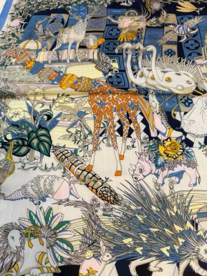 silkesscarf med djurmönster - Färgstark silkesscarf med detaljerat djurmönster i blått, orange, gult och vitt. Scarfen har motiv av giraffer, flodhästar, fåglar och blommor mot en bakgrund av geometriska former och resväskor. Perfekt accessoar för att lyfta din outfit. 