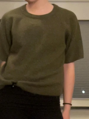 Olivgrön stickad t-shirt - Säljer en olivgrön stickad t-shirt med korta ärmar och rund hals. Tröjan har en enkel, avslappnad passform och ribbstickade kanter vid ärmslut och nederkant. Perfekt för lager på lager och en chill look.