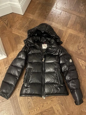 Svart dunjacka från Moncler - Snygg svart dunjacka från Moncler med glansig finish och quiltad design. Jackan har en stor huva, två dragkedjeförsedda fickor och Moncler-logga på ärmen. Perfekt för kalla vinterdagar och riktigt trendig stil.