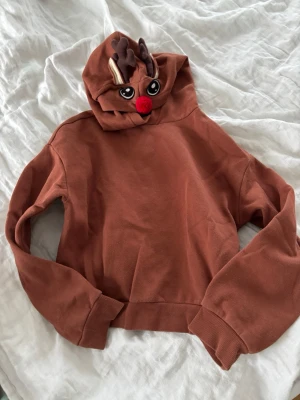 Brun hoodie med renmotiv och luva - Mysig brun hoodie med luva som har ett gulligt renansikte, röda nosen och mjuka horn.🦌🎄 Perfekt för dig som vill sticka ut med en julig och lekfull stil. Tillverkad i mjuk bomullsblandning.✨❤️💚