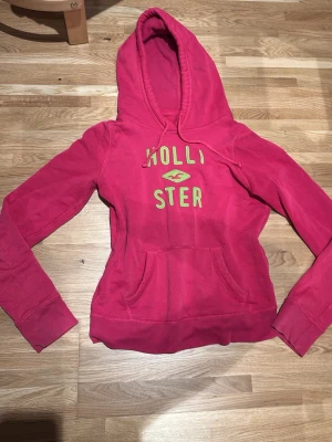 Rosa hoodie från Hollister - Säljer en snygg rosa hoodie från Hollister med limegröna bokstäver och logga på bröstet. Hoodien har huva med dragsko och en stor magficka.is Tillverkad i mjukt bomullsmaterial och har långa ärmar med ribbade muddar.