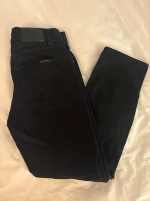 Svarta jeans från Nudie Jeans - Säljer ett par svarta jeans från Nudie Jeans med klassisk femficksmodell och diskret vågmönster på bakfickan. Jeansen har en svart läderpatch i midjan och är tillverkade i kraftig denim. Perfekta för en clean och stilren look.