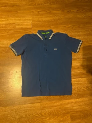 Blå piké från BOSS med vit kant - Snygg blå pikétröja från BOSS med vit och grå rand på krage och ärmslut. Klassisk passform med korta ärmar och knäppning framtill. Perfekt för en stilren och avslappnad look.