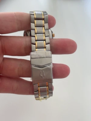 Bulova klocka i stål och guld - Stilren Bulova klocka med armband i rostfritt stål och guldfärgade detaljer. Urtavlan är vit med guldfärgade visare och index samt datumvisning. Klassisk design med tvåfärgat länkarmband och Bulova-logga på spännet.