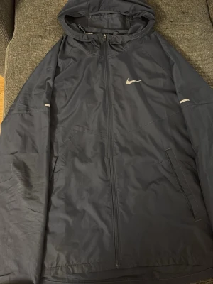 Svart Nike vindjacka med huva - Svart vindjacka från Nike med huva och hel dragkedja framtill. Jackan har två sidofickor med dragkedja och reflekterande detaljer på ärmarna. Perfekt för löpning eller utomhusaktiviteter. Lätt och smidig design med Nike-logga på bröstet.