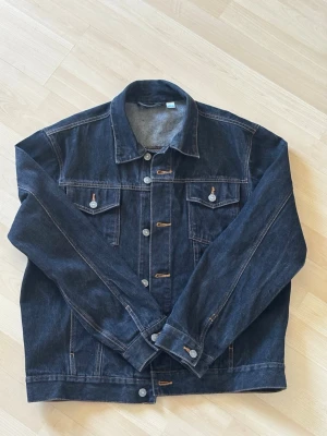Mörkblå jeansjacka från Real Men - Snygg mörkblå jeansjacka från Real Men med klassisk rak passform. Jackan har två bröstfickor med lock och knapp, samt knappar framtill och vid ärmsluten. Tillverkad i slitstarkt denimtyg med kontrastsömmar för en tidlös look. (Passer till storlek S M 