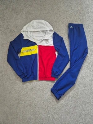 Lacoste - Säljer ett riktigt snyggt tvådelat sportset från Lacoste med jacka och byxor. Jackan har huva och dragkedja, och är blå, röd, vit och gul med stor Lacoste-logga och krokodil på bröstet. Byxorna är blå med resår i midja och benslut. Perfekt för träning eller chill.