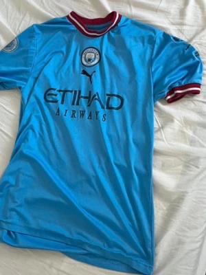 Manchester City matchtröja Haaland #9 - Säljer en ljusblå Manchester City matchtröja från Puma med Haaland och nummer 9 på ryggen. Tröjan har klubbmärke på bröstet, sponsortryck och röda/vita detaljer vid krage och ärmslut. Perfekt för fotbollsfans som vill visa sitt stöd på läktaren eller planen.