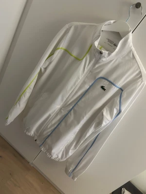 Vit tracksuit från Lacoste - Snygg vit vindjacka och byxor från Lacoste med dragkedja framtill. Jackan har tunna blå och gula detaljer längs ärmar och bröst, samt klassisk Lacoste-logga på vänster sida samt gäller byxorna det är tillverkad i lätt och ventilerande material, perfekt för sport eller träning. Elastiska muddar vid ärmslut och nederkant.