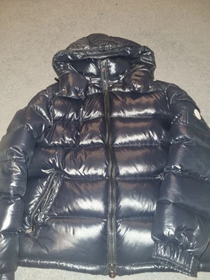 Mörkblå pufferjacka från Moncler - Snygg mörkblå pufferjacka från Moncler med glansig finish, stor huva och dragkedjor på fickorna. Klassisk Moncler-logga på ärmen och quiltad design som håller värmen. Perfekt för kalla vinterdagar och riktigt trendig look. Storlek 3/L men passar mer som M.