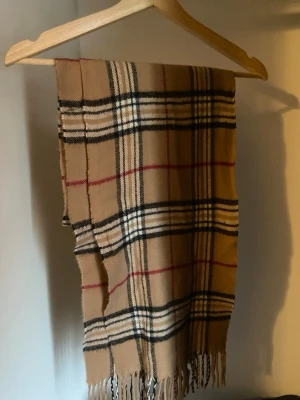 Burberry HalsDuk  - Klassisk beige halsduk med rutigt mönster i svart, vitt och rött. Halsduken har fransar i ändarna och är tillverkad i ett mjukt material som känns skönt mot huden. Perfekt accessoar för att lyfta din outfit med en tidlös stil.