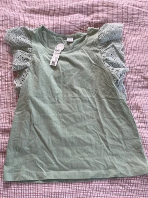Grön topp med volangärmar och spets - Superfin ljusgrön topp från Lindex med volangärmar i broderad spets. Ärmlös modell med rund halsringning och mjukt bomullstyg. Perfekt för dig som vill ha en söt och trendig look med extra detaljer på axlarna. I storlek 158/164. Ny med prislapp. 