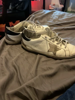 Golden Goose vita sneakers med stjärna - Säljer ett par vita Golden Goose sneakers med grå stjärna på sidan och svarta detaljer vid hälen. Skorna har låga skaft, grå snören och är tillverkade i läder. Klassisk streetstil med slitna detaljer och ikonisk design.