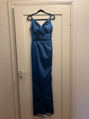 Blå satinklänning med v-ringning - Elegant blå långklänning med v-ringad omlott framtill och smala axelband. Klänningen har en markerad midja och är tillverkad i ett glansigt material som ger en lyxig känsla. Perfekt för dig som vill sticka ut med en stilren och modern look.