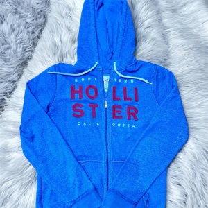 Blå zip hoodie från Hollister - Blå hoodie från Hollister med dragkedja och stor huva. Framsidan har stora röda bokstäver med texten 'Hollister California'. Mjukt material och vita dragsnören vid huvan. Perfekt för en avslappnad och cool stil.                                      Hör av er vid eventuella frågor!