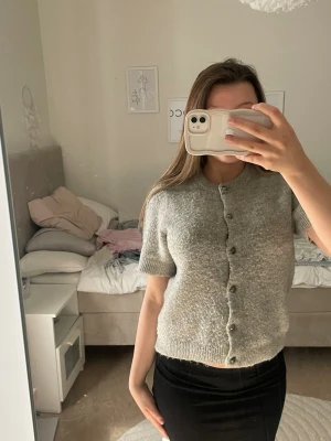 Grå kortärmad kofta med knappar - Superfin grå kofta med korta ärmar och rund hals. Koftan har en rak passform och knäpps med små knappar framtill. Perfekt att styla över en topp eller klänning. Mjuk och skön, troligtvis i ull- eller mohairblandning.