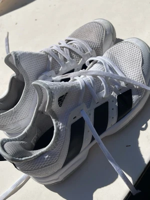 Adidas vita sneakers med svarta ränder - Fräscha vita sneakers från Adidas med de klassiska tre svarta ränderna på sidorna. Ovandelen är i ventilerande mesh och syntetmaterial, med vit sula och snörning. Perfekta för dig som gillar sportig och stilren look.