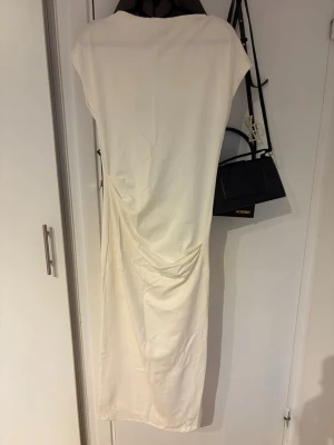Lång vit klänning med drapering - Snygg långklänning i vit färg med draperad detalj på sidan. Klänningen har en enkel och stilren design med korta ärmar och rak passform. Perfekt för dig som gillar minimalistisk stil och vill ha något elegant till sommaren.