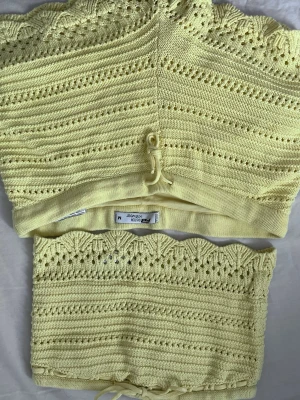 Gul stickad topp från FJ Sista Knitwear - Söt ljusgul stickad topp med virkade detaljer och knytband nertill. Toppen har ett fint hålmönster och dekorativa små stenar framtill. Perfekt för dig som gillar unika och somriga plagg.