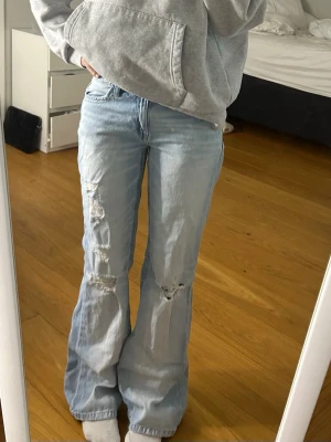 Gina jeans  - Säljer ett par ljusblå bootcut jeans med slitningar och hål på båda benen. Jeansen har hög midja och klassisk femficksdesign. Perfekta för en avslappnad och trendig look. Materialet är denim i bomull.