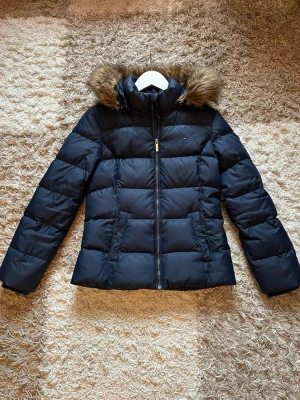 Marinblå pufferjacka med fuskpäls från Tommy Hilfiger - Snygg svart pufferjacka från Tommy Hilfiger med avtagbar huva och fuskpäls. Jackan har dragkedja framtill, två fickor med dragkedja och quiltad design. Perfekt för kalla dagar och ger en stilren look.