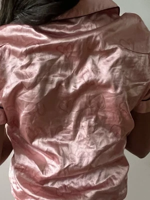 Rosa satinpyjamas med hjärtmönster - Söt pyjamas i glansigt rosa satintyg med diskret hjärtmönster. Kortärmad skjorta med svarta kontrastkanter, bröstficka och knappar framtill. Matchande shorts. Perfekt för mysiga kvällar hemma.