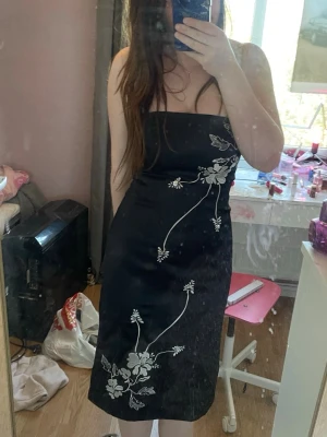 Svart axelbandslös klänning med blommor - Snygg svart axelbandslös klänning i satin med vita broderade blommor framtill. Klänningen har en smickrande slim passform och slutar strax under knät. Perfekt för dig som vill ha en elegant look med fina detaljer.