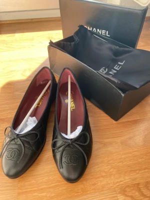 Svarta quiltade ballerinaskor Chanel - Snygga svarta ballerinaskor från Chanel i quiltat läder med rosett framtill och klassisk CC-logga på tån. Insidan är vinröd och skon har en elegant, tidlös form. Skorna levereras med box och dustbag. Pris kan diskuteras!💕