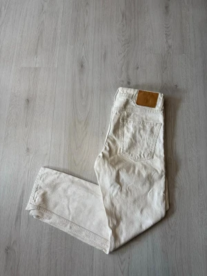 Jack & Jones Jeans - Säljer ett par vita jeans med rak passform och knappgylf. Jeansen har klassiska fem fickor och en läderpatch bak i midjan. Perfekta för en clean och stilren look. Materialet är denim i bomull. Möjligen en liten fläck på någon av benen