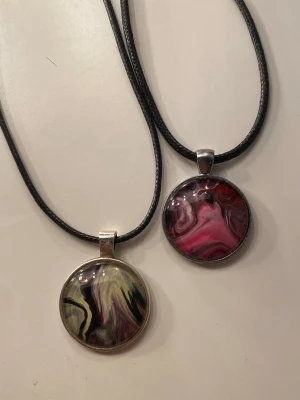Två runda halsband med marmorhänge - Säljer två snygga halsband med runda hängen i marmorliknande mönster. Ena hänget har rosa och svarta toner, det andra har grönt, svart och lila. Båda har svarta snören och silverfärgade detaljer. Helt oanvända! 10kr styck eller båda för 17kr! 🫶