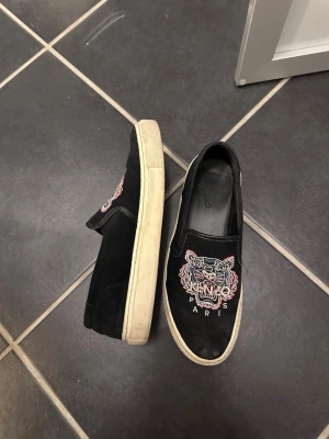 Svarta Kenzo slip-on sneakers - Svarta slip-on sneakers från Kenzo med ikoniskt broderat tigerhuvud och texten 'Paris' framtill. Skorna har vit sula och är tillverkade i mocka med elastiska paneler på sidorna för enkel påtagning. Perfekta för en streetwear-look.