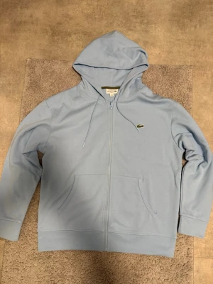 Ljusblå zip hoodie från Lacoste - Snygg ljusblå hoodie från Lacoste med dragkedja framtill och klassisk krokodil-logga på bröstet. Hoodien har två stora fickor fram, justerbar huva med snören och ribbade muddar. Perfekt för en chill och stilren look.