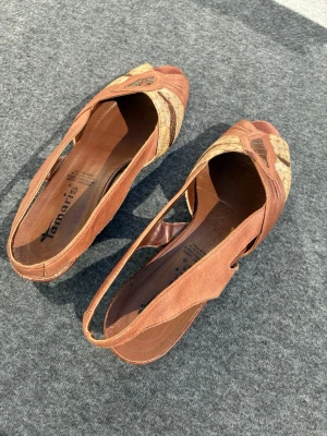 Bruna peep-toe pumps från Tamaris - Snygga bruna pumps med låg klack från Tamaris med öppen tå och slingback-rem. Skorna har ett coolt mönster i beige och brunt på ovandelen och är gjorda i läder. Perfekta för dig som gillar retrovibbar och unika detaljer.