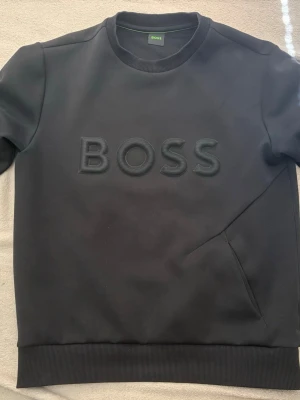 Svart sweatshirt från BOSS - Snygg svart sweatshirt från BOSS med stort broderat logotyptryck på bröstet. Tröjan har rund halsringning, ribbade muddar vid ärmslut och nederkant samt en stilren design. Perfekt för en clean och modern look.