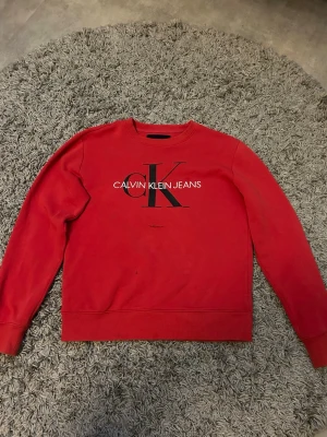 Röd sweatshirt från Calvin Klein Jeans - Röd sweatshirt med rund hals från Calvin Klein Jeans. Stor svart CK-logga och vit text framtill. Bra skick