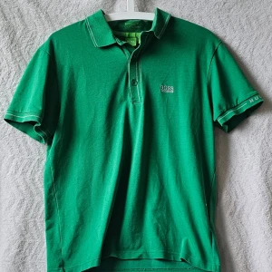 Grön pikétröja från Hugo Boss - Snygg grön pikétröja från Hugo Boss med klassisk krage och knäppning framtill. Tröjan har korta ärmar och vit kontrastsöm vid krage och ärmslut. Boss-logga broderad på bröstet. Perfekt för en stilren look.